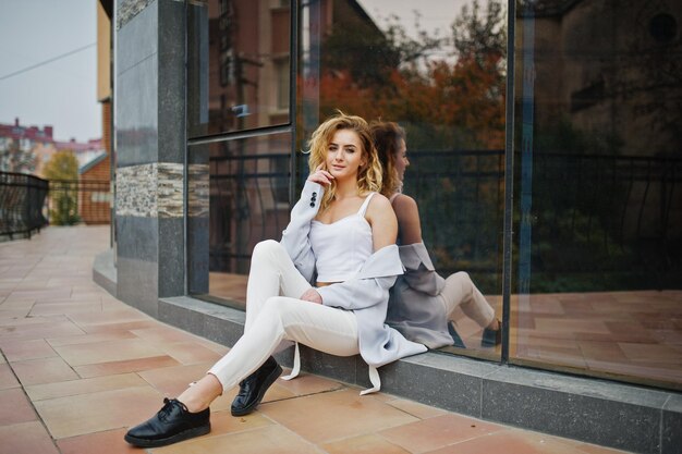 Elegante modelo rubia rizada vestida de blanco posando contra una ventana grande