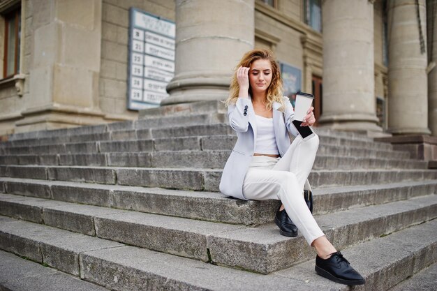 Elegante modelo rubia rizada vestida de blanco con una taza de café posando en las escaleras al aire libre