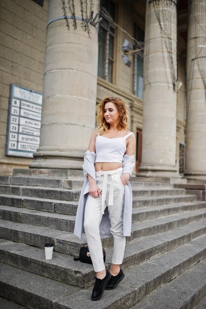 Elegante modelo rubia rizada vestida de blanco con una taza de café posando en las escaleras al aire libre