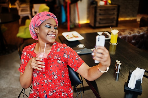 Elegante mujer africana con camisa roja y sombrero posó en un café interior bebiendo limonada de fresa y haciendo selfie por teléfono