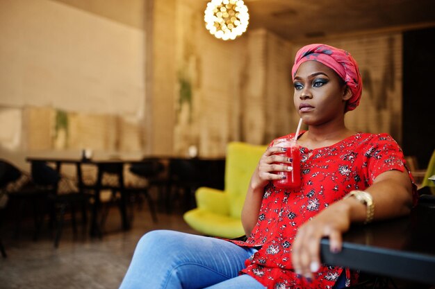 Elegante mujer africana con camisa roja y sombrero posó en un café interior y bebiendo limonada de fresa