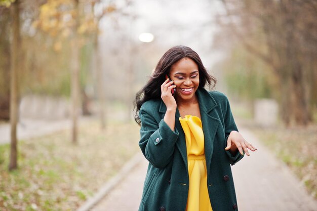 Elegante mujer afroamericana con abrigo verde y vestido amarillo posada contra el parque de otoño y hablando por teléfono