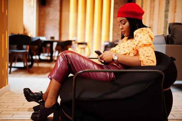 Elegante mujer afroamericana en boina francesa roja blusa de lunares con cadena de cuello dorado grande y pantalones de cuero sentada en el puf con teléfono móvil