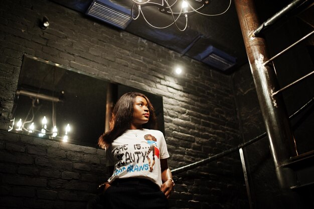 Elegante mujer afroamericana en camiseta impresa posó escaleras de acero interiores
