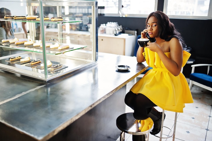 Foto gratuito elegante mujer afroamericana en vestido amarillo posó en el café con una taza de capuchino y hablando por teléfono