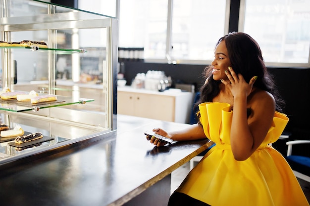 Elegante mujer afroamericana en vestido amarillo posó en el café con teléfono móvil a mano