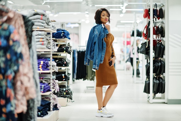 Elegante mujer afroamericana con vestido de túnica marrón y chaqueta de jeans posada en la tienda de ropa Es hora de ir de compras