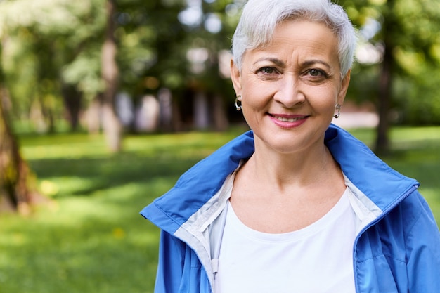 Elegante mujer alegre de jubilación con un agradable paseo por el bosque soleado en un día de verano con una sonrisa feliz, disfrutando de un clima hermoso y aire fresco, árboles y pasto verde