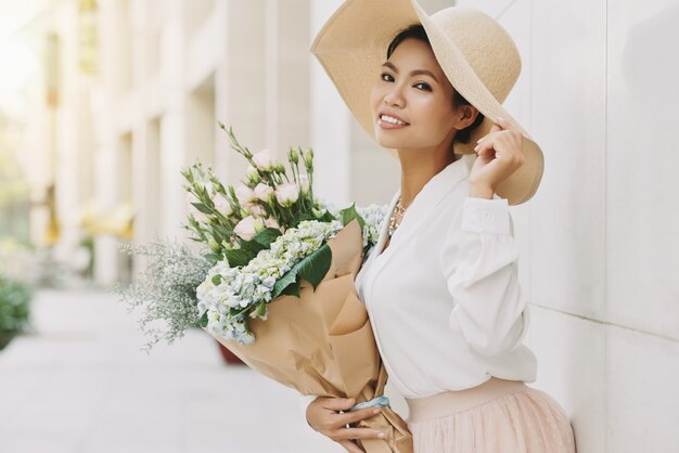 Elegante mujer asiática bien vestida en gran sombrero para el sol posando en la calle urbana con flores.