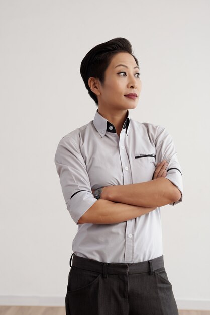 Elegante mujer asiática en pantalones y camisa posando en estudio con brazos cruzados