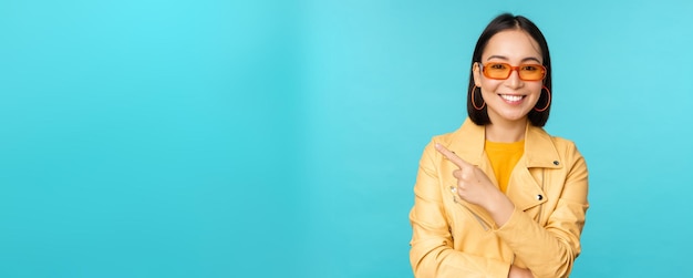 Foto gratuita elegante mujer asiática sonriente con gafas de sol apuntando con el dedo a la izquierda mostrando una pancarta publicitaria de pie con un moderno abrigo amarillo sobre fondo azul.