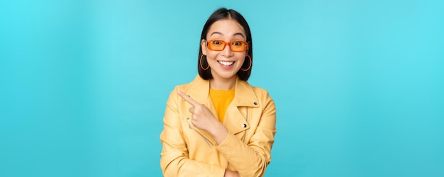 Elegante mujer asiática sonriente con gafas de sol apuntando con el dedo a la izquierda mostrando una pancarta publicitaria de pie con un moderno abrigo amarillo sobre fondo azul.