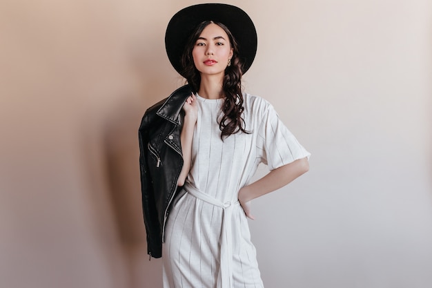 Elegante mujer asiática en vestido blanco mirando a cámara. Mujer China confía en sombrero con chaqueta de cuero.