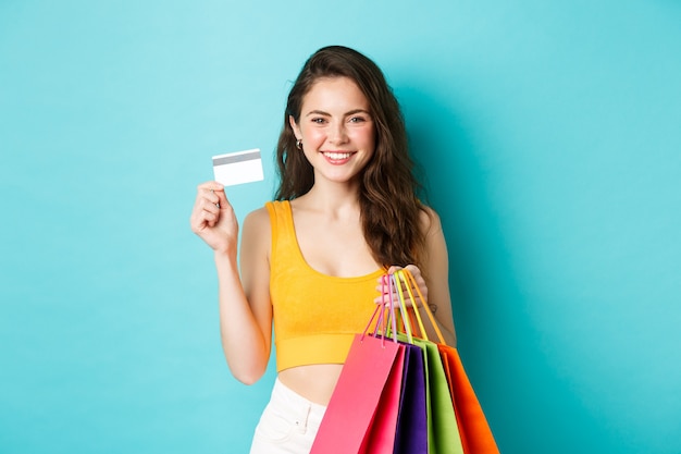 Elegante mujer atractiva que muestra la tarjeta de crédito de plástico que solía pagar por la ropa nueva, sosteniendo bolsas de la compra con productos, de pie sobre fondo azul.