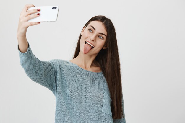 Elegante mujer atractiva sonriendo mostrando la lengua, tomando selfie