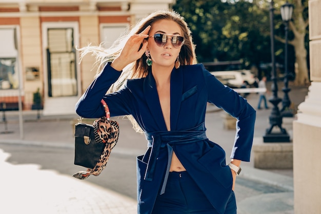 Elegante mujer atractiva vestida con elegante traje azul y gafas de sol caminando en la calle sosteniendo el bolso