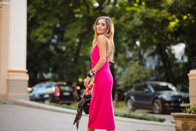 Elegante mujer atractiva con vestido de verano sexy rosa caminando en la calle sosteniendo el bolso