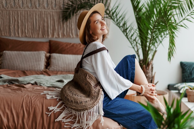 Elegante mujer bonita con sombrero de paja y blusa blanca posando en casa, sentado en la cama