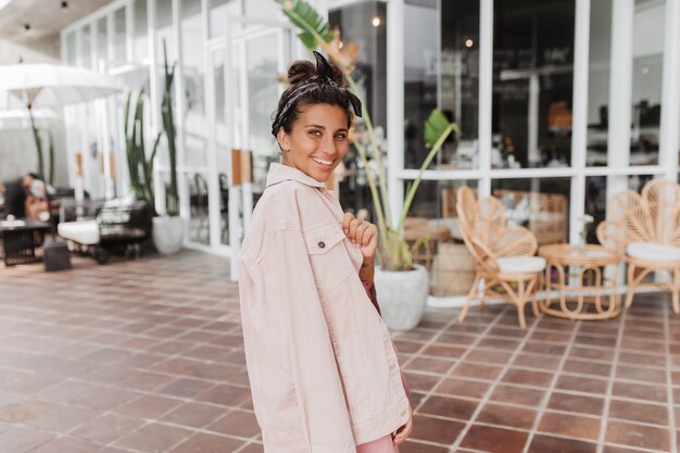 Elegante mujer bronceada en diadema y chaqueta de mezclilla rosa posando contra street cafe