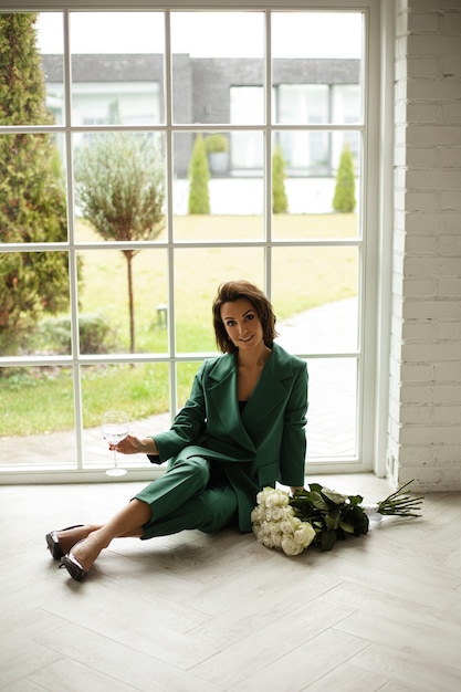 Elegante mujer caucásica con cabello oscuro en traje verde posa para la cámara cerca del gran bouquiet de flores blancas y sonrisas