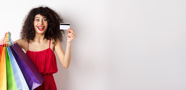 Foto gratuita elegante mujer caucásica con cabello rizado y vestido rojo mostrando bolsas de compras y su crédito de plástico c