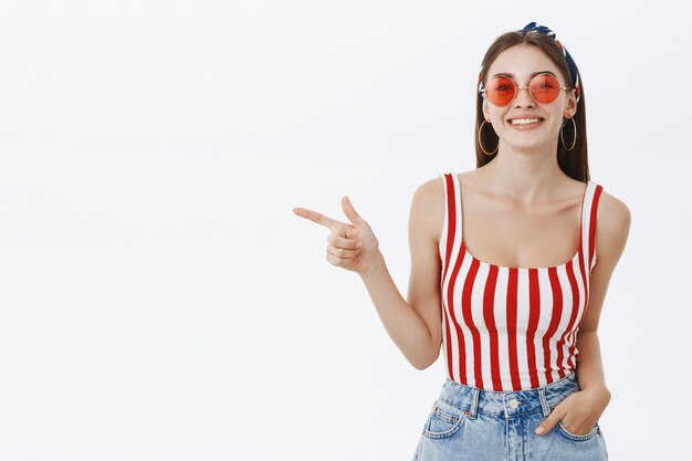 Elegante mujer caucásica confiada en gafas de sol redondas de moda y top rayado sosteniendo la mano en el bolsillo apuntando a la izquierda con el dedo índice y sonriendo amable y complacido