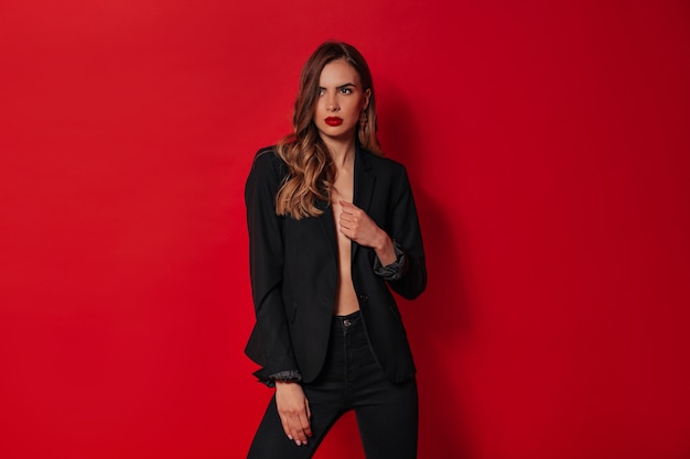 Foto gratuita elegante mujer confiada en traje negro posando sobre pared roja