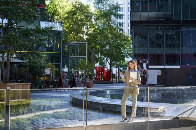 Elegante mujer corporativa en traje de pie con una computadora portátil y documentos de trabajo usando un teléfono móvil mirando a