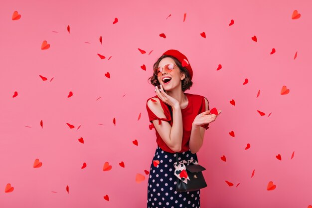 Elegante mujer despreocupada posando en el día de San Valentín. Riendo a chica rizada glamorosa en boina de pie bajo confeti rojo.