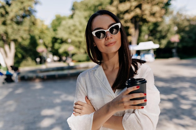 Elegante mujer elegante con cabello oscuro y liso con gafas de sol y camisa blanca sostiene una taza con café y posa a la cámara a la luz del sol en un parque verde de verano en la ciudad