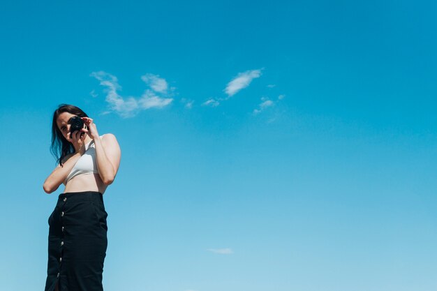 Elegante mujer fotógrafa tomando foto con cámara contra el cielo azul