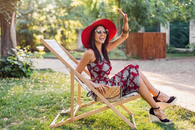 Elegante mujer hermosa sentada en la tumbona con traje de estilo tropical, agitando la mano, tendencia de la moda de verano, bolso de paja, sombrero rojo, gafas de sol, accesorios, sonriente, feliz humor, vacaciones