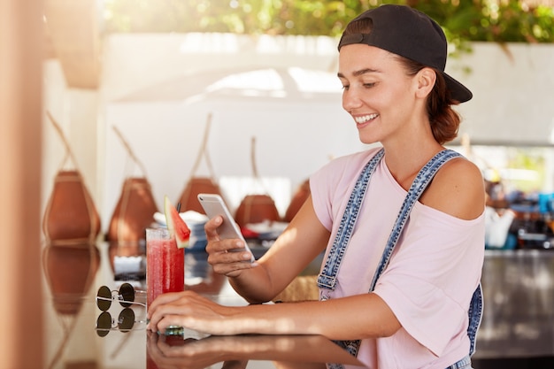 Elegante mujer hipster con gorra, que se alegra de recibir mensajes de texto en el teléfono móvil, navega por internet en un acogedor café durante la recreación de verano, bebe batido, viste ropa de moda. Gente y estilo