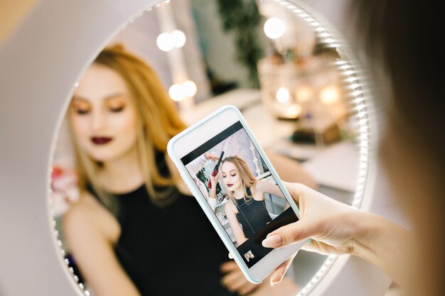 Elegante mujer joven y bonita haciendo fotos en el teléfono en el espejo durante la realización de peinado en la peluquería. Modelo de moda elegante, preparándose para la fiesta, celebración, look de lujo