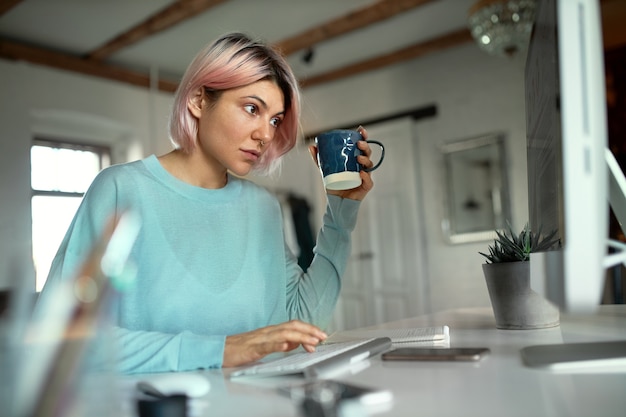 Elegante mujer joven con cabello rosado sentada en su lugar de trabajo escribiendo en el teclado bebiendo té usando