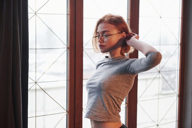 Elegante mujer joven. Chica rubia sexy en gafas, ropa interior y sin sujetador debajo de la camisa posando cerca de la ventana de la habitación