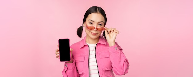 Elegante mujer joven coreana con gafas de sol que muestra la interfaz de la aplicación del teléfono móvil de la pantalla del teléfono inteligente o el sitio web de pie sobre fondo rosa