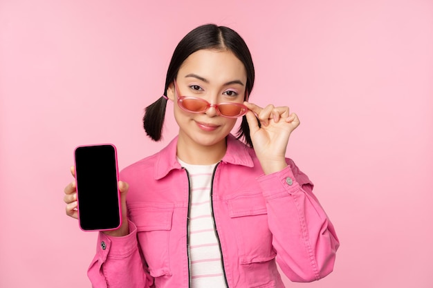 Elegante mujer joven coreana con gafas de sol que muestra la interfaz de la aplicación del teléfono móvil de la pantalla del teléfono inteligente o el sitio web de pie sobre fondo rosa