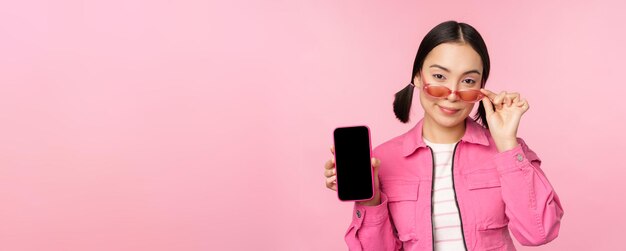 Elegante mujer joven coreana con gafas de sol que muestra la interfaz de la aplicación del teléfono móvil de la pantalla del teléfono inteligente o el sitio web de pie sobre fondo rosa