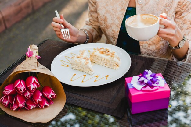 Elegante mujer joven sentada en el café, sosteniendo una taza de capuchino y comiendo un sabroso pastel,