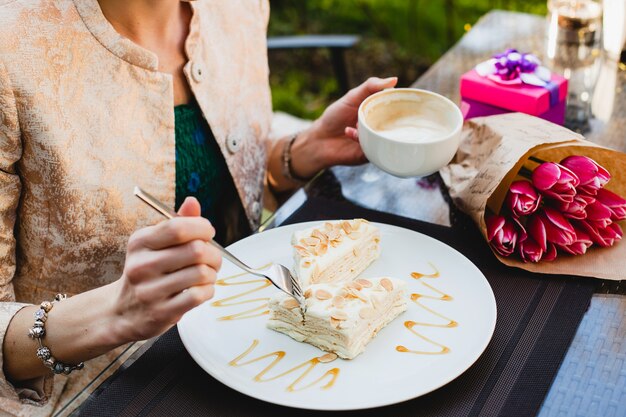 Elegante mujer joven sentada en el café, sosteniendo una taza de capuchino y comiendo un sabroso pastel,