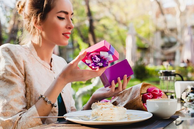 Elegante mujer joven sentada en la cafetería, sosteniendo el cuadro actual, sonriendo