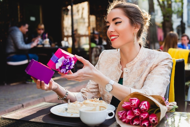 Elegante mujer joven sentada en la cafetería, sosteniendo el cuadro actual, sonriendo