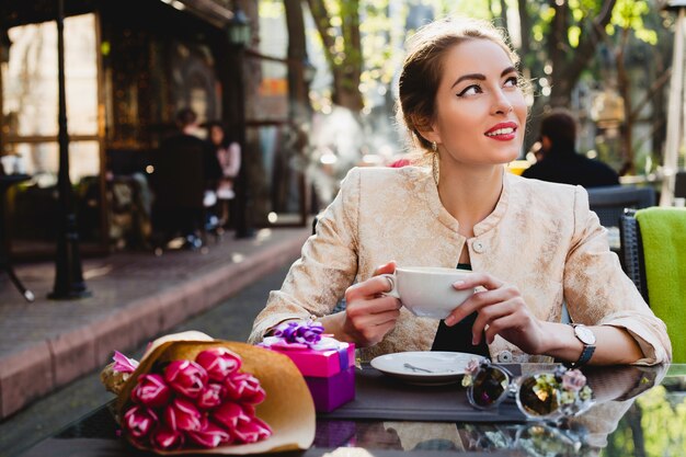 Elegante mujer joven sentada en la cafetería, sosteniendo una taza de capuchino