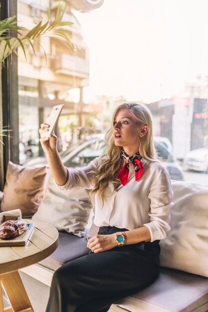 Elegante mujer joven sentada en la cafetería tomando selfie desde un teléfono inteligente