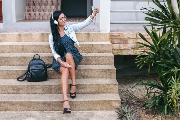 Elegante mujer joven sentada en las escaleras con el teléfono inteligente, escuchando música en auriculares, tomando una foto