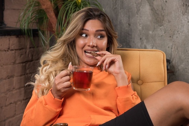 Elegante mujer joven sentada en un sillón sosteniendo una taza de té y comiendo galletas.