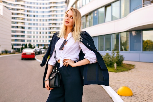 Elegante mujer joven con traje azul marino de moda, posando cerca de edificios modernos, accesorios de moda, sonriendo al final disfrutando de un día soleado de verano, caminando cerca de la oficina.