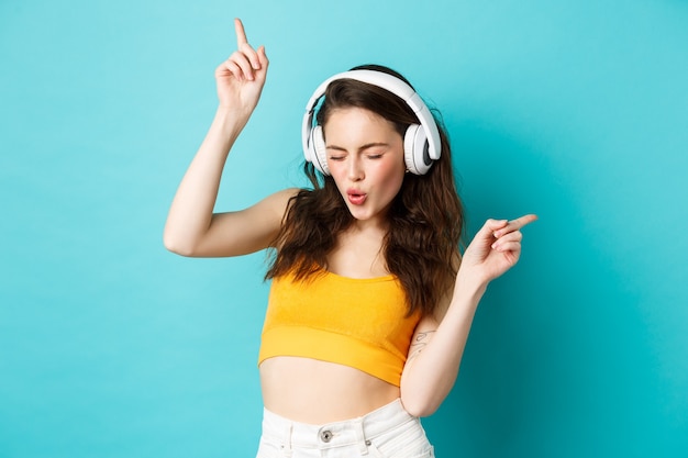 Elegante mujer moderna en auriculares, escuchando música y bailando, disfrutando de sus canciones favoritas en auriculares, de pie contra el fondo azul.