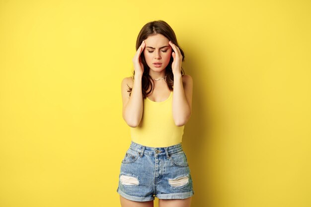 Elegante mujer moderna frotando las sienes, con dolor de cabeza, migraña, frunciendo el ceño y haciendo muecas de dolor en la cabeza, de pie contra el fondo amarillo